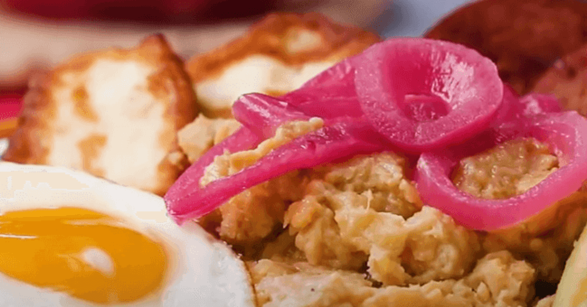 A close-up of a breakfast plate featuring fried egg, golden-brown fried cheese, mashed plantains, and pickled red onions.